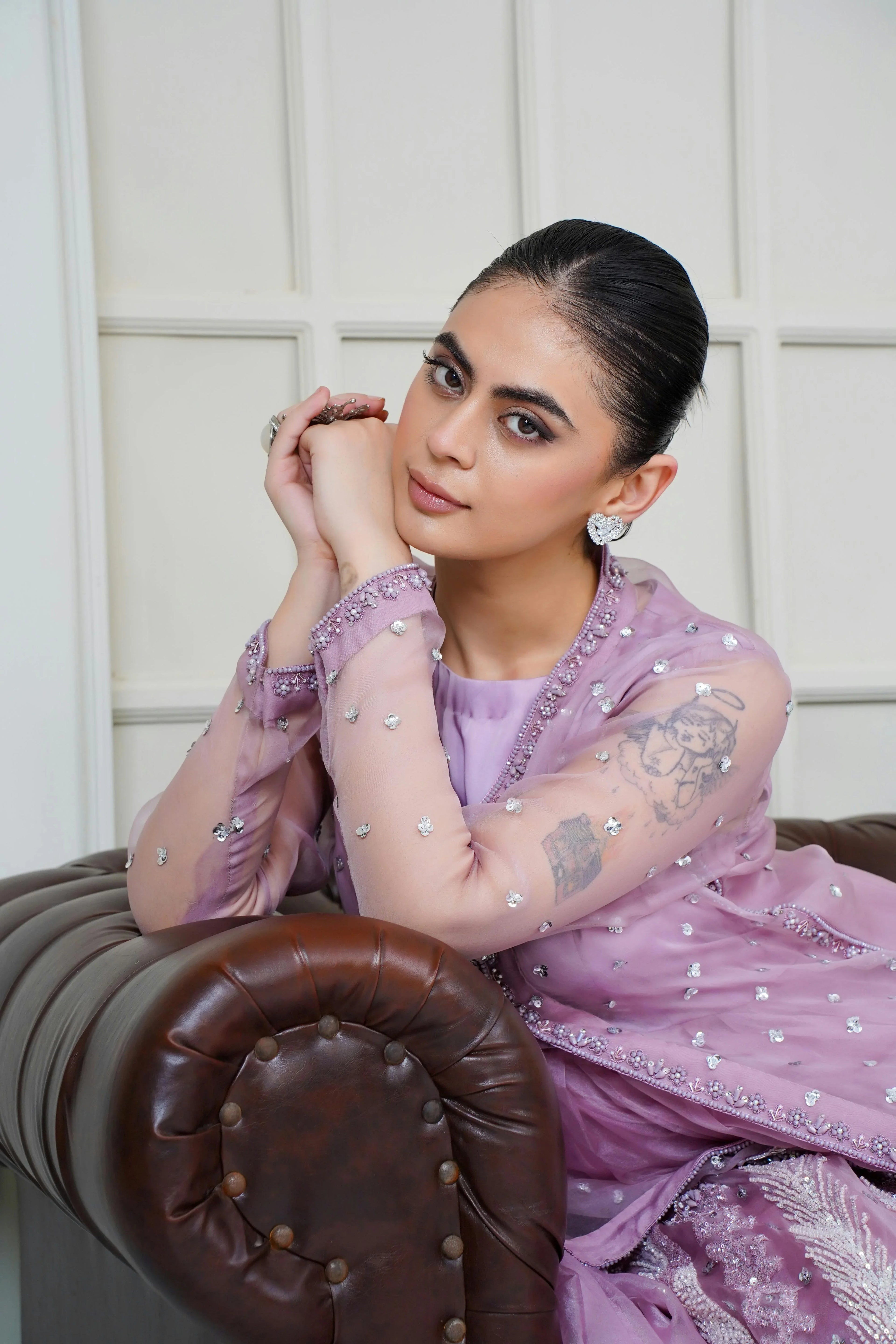 Woman in lilac embroidered Pakistani formal wear with sheer sleeves, sitting on a sofa.