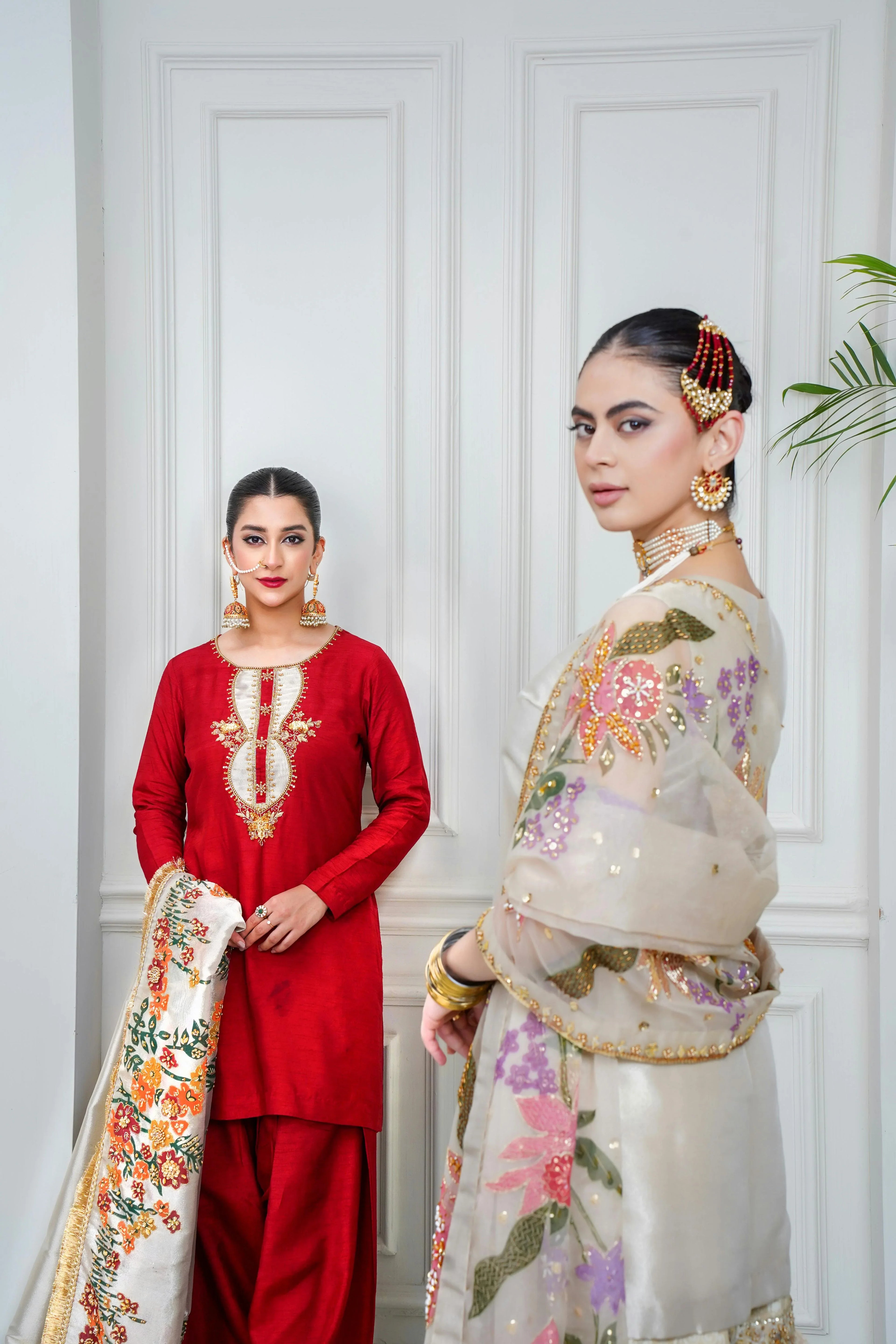 Two women in elegant Pakistani bridal and formal wear, red embroidered dress and ivory festive suit.