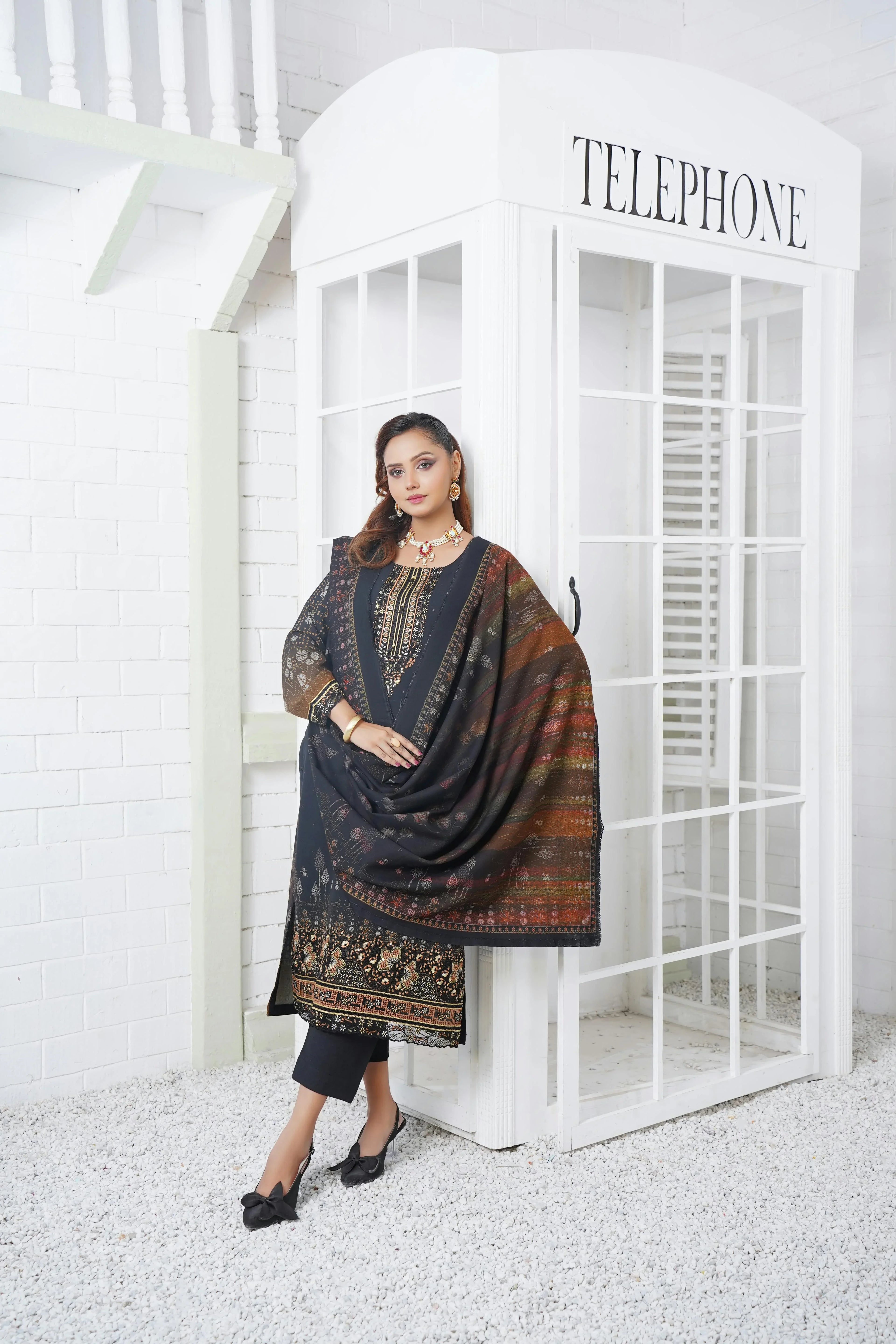 Woman in elegant black Pakistani dress with embroidered lawn, festive wear Pakistan, designer suit Pakistan, standing by white telephone booth.